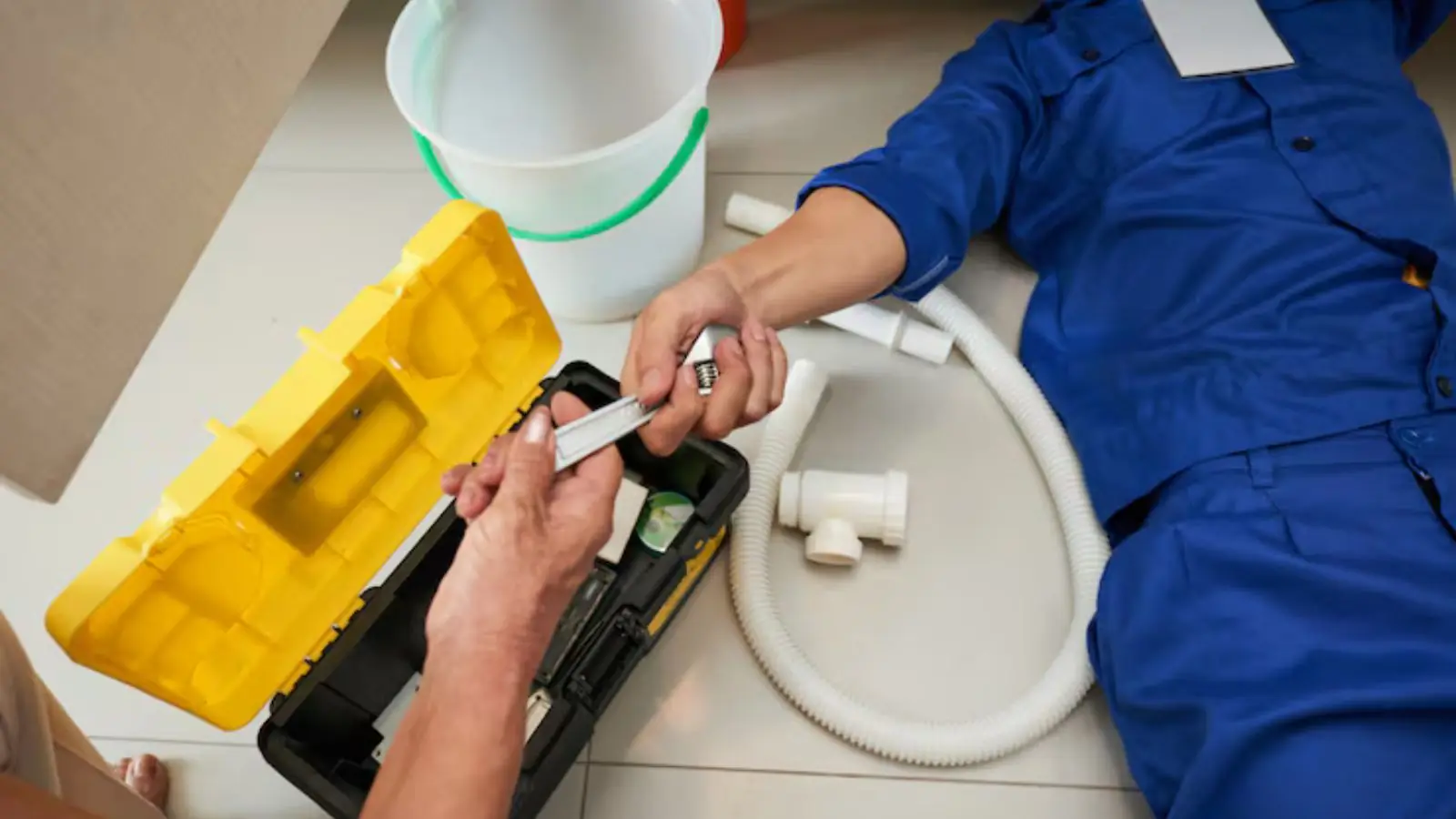 Hands working on a white drain hose beside a yellow toolbox during a washing machine hose repair