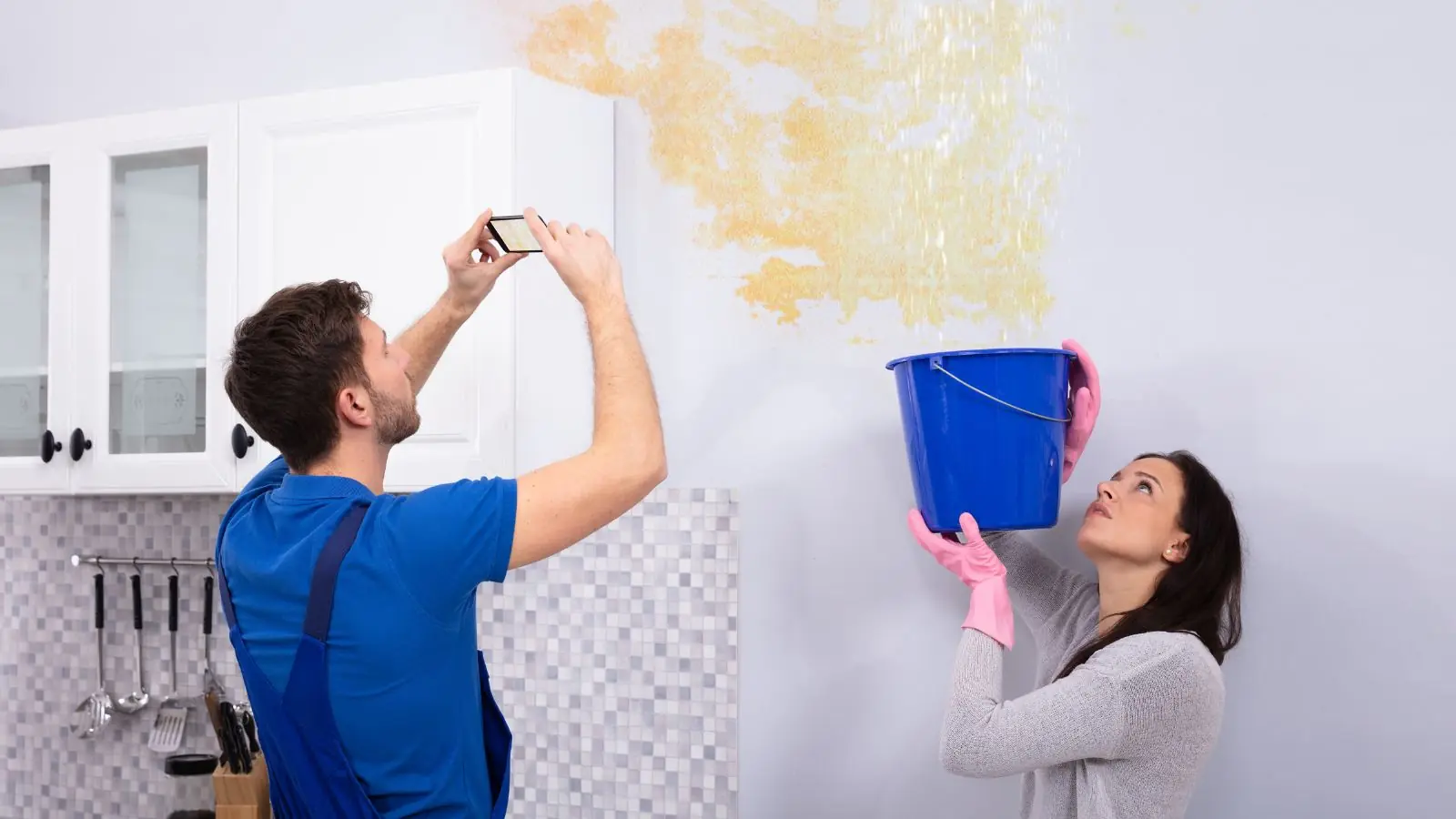 Homeowner photographing a water stain on a wall while another holds a bucket, documenting damage for an insurance claim
